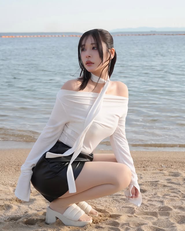 一名女性坐在沙灘上，穿著白色露肩上衣和黑色短裙，旁邊是平靜的海面和藍天。