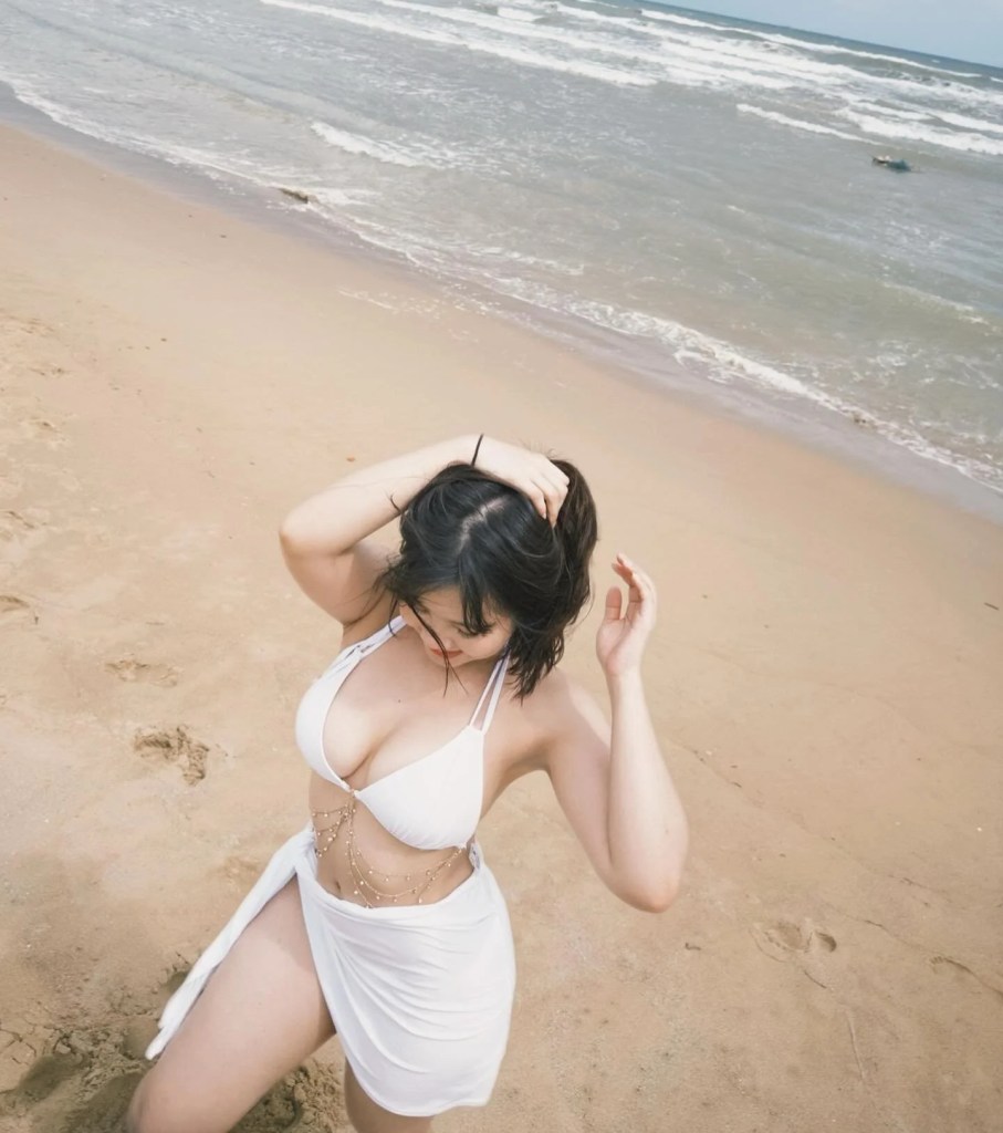在沙灘上，一名女性穿著白色比基尼和輕薄裙子，正撥弄頭髮，海浪在背景中拍打著海岸。