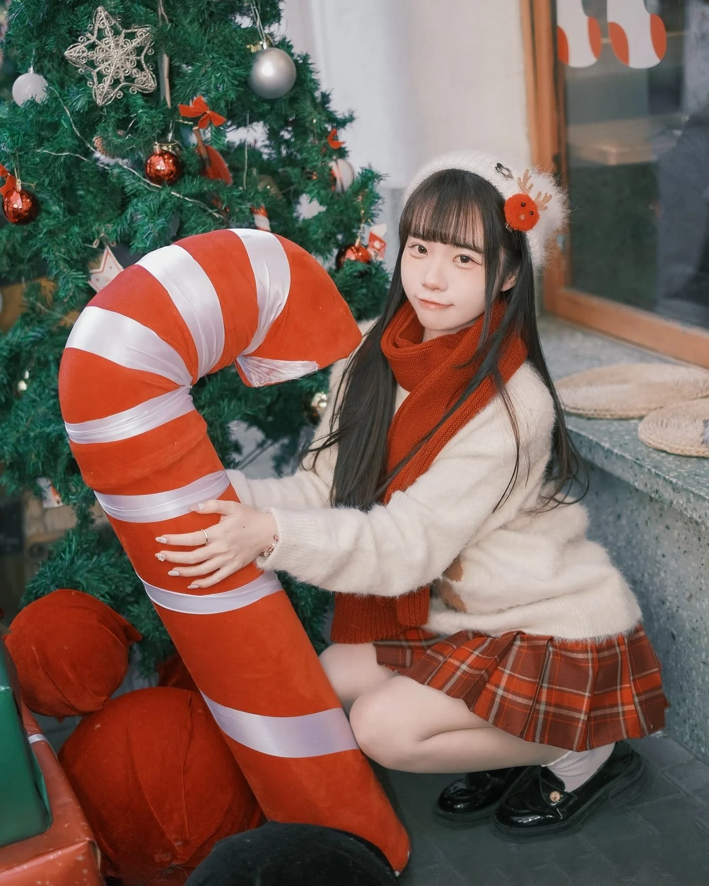 一名女孩坐在聖誕樹旁，抱著一根紅白相間的甜筒，穿著毛衣和格子裙，頭戴裝飾的帽子，表情可愛。