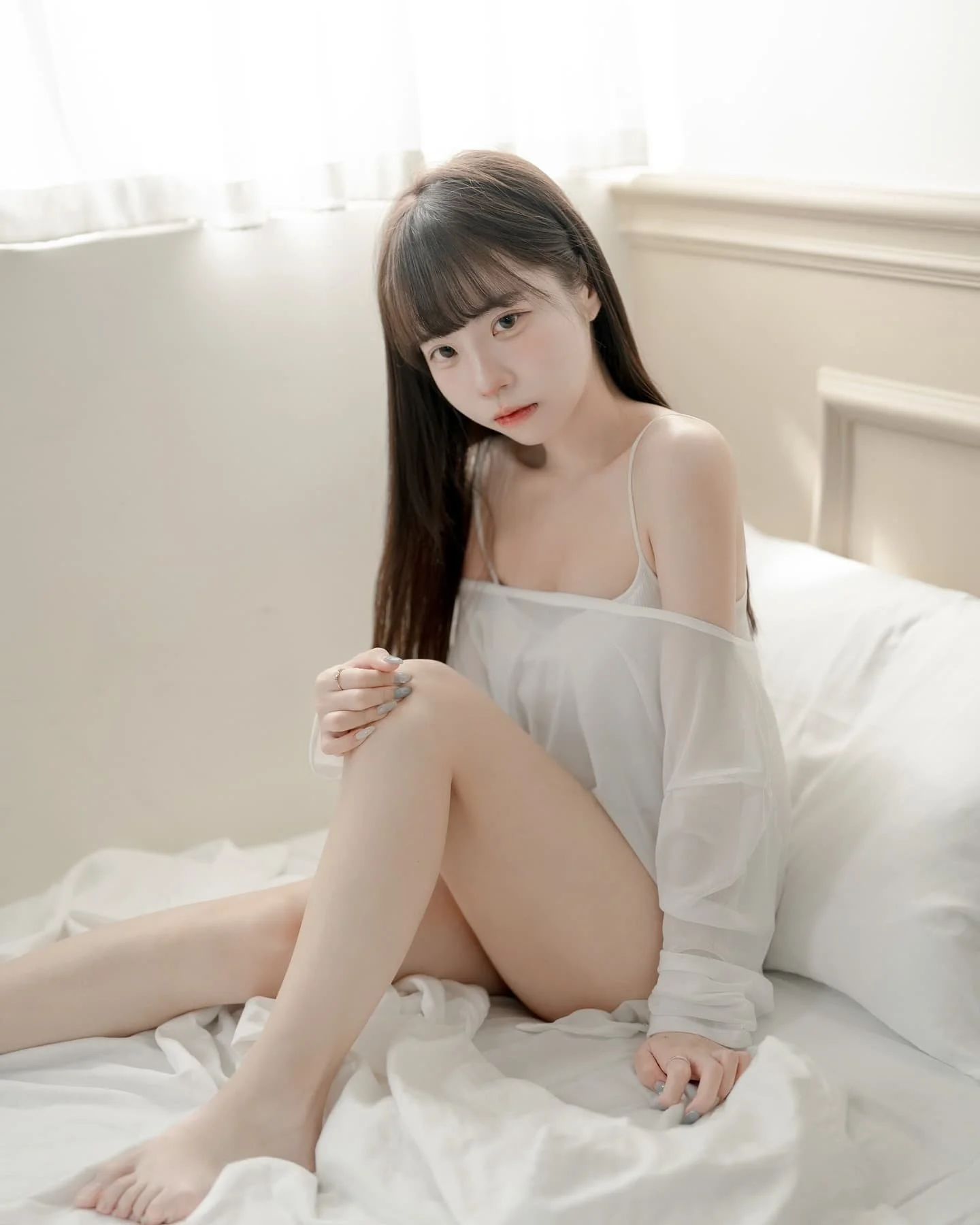 一位女性坐在床上，身穿輕薄的白色上衣，表情柔和，背景有自然光透入。