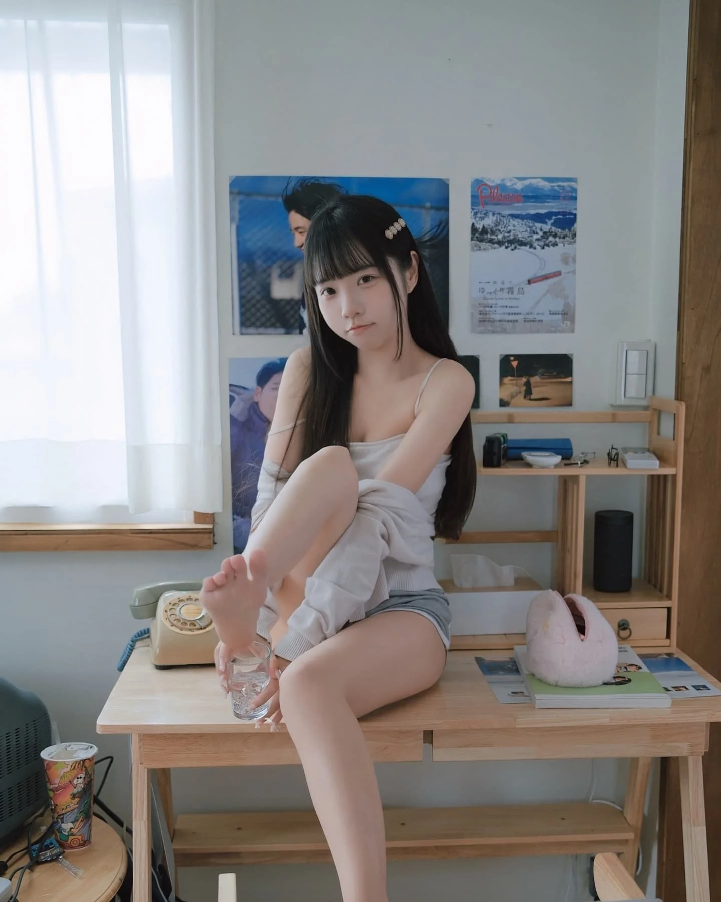 一位女孩坐在桌子上，穿著淺色上衣和短褲，手中拿著一杯水，展現出悠閒的居家環境。背景牆上有幾張海報，桌面上擺放著電話和其他物品。