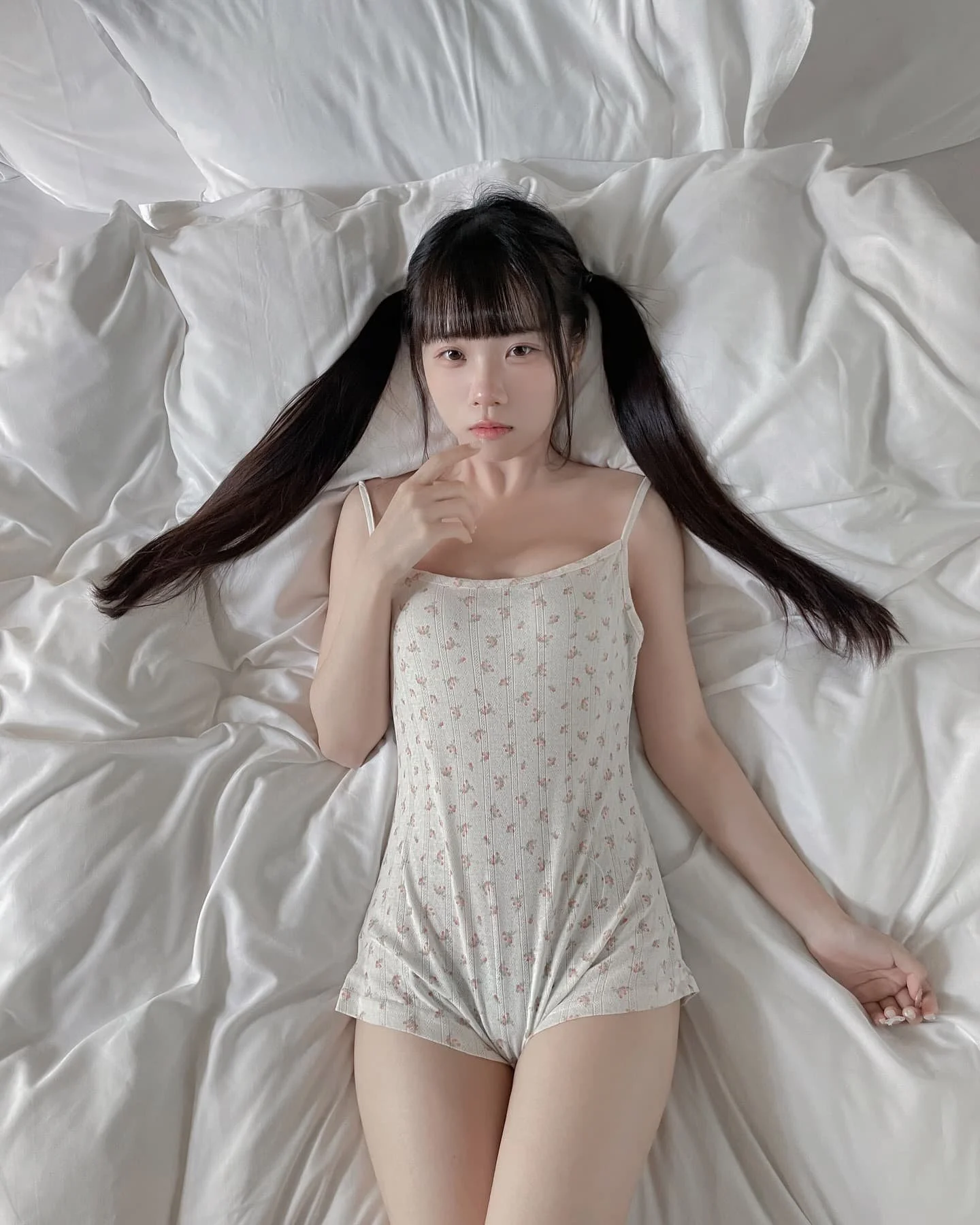 一位女性穿著輕薄的睡衣，躺在白色床單上，長髮披肩，手指輕觸嘴唇，表情凝思。