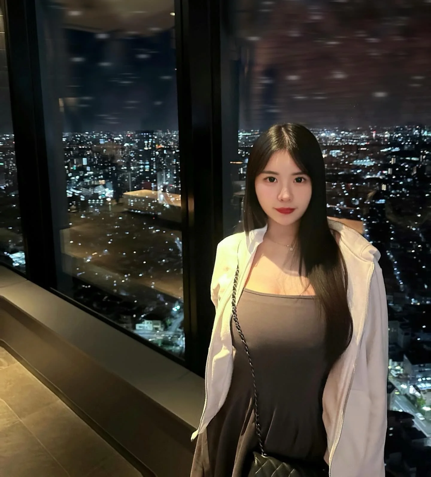 一位穿著灰色連衣裙的年輕女性，站在高樓的窗邊，背後是璀璨的城市夜景，窗外可見燈火通明的城市景觀。