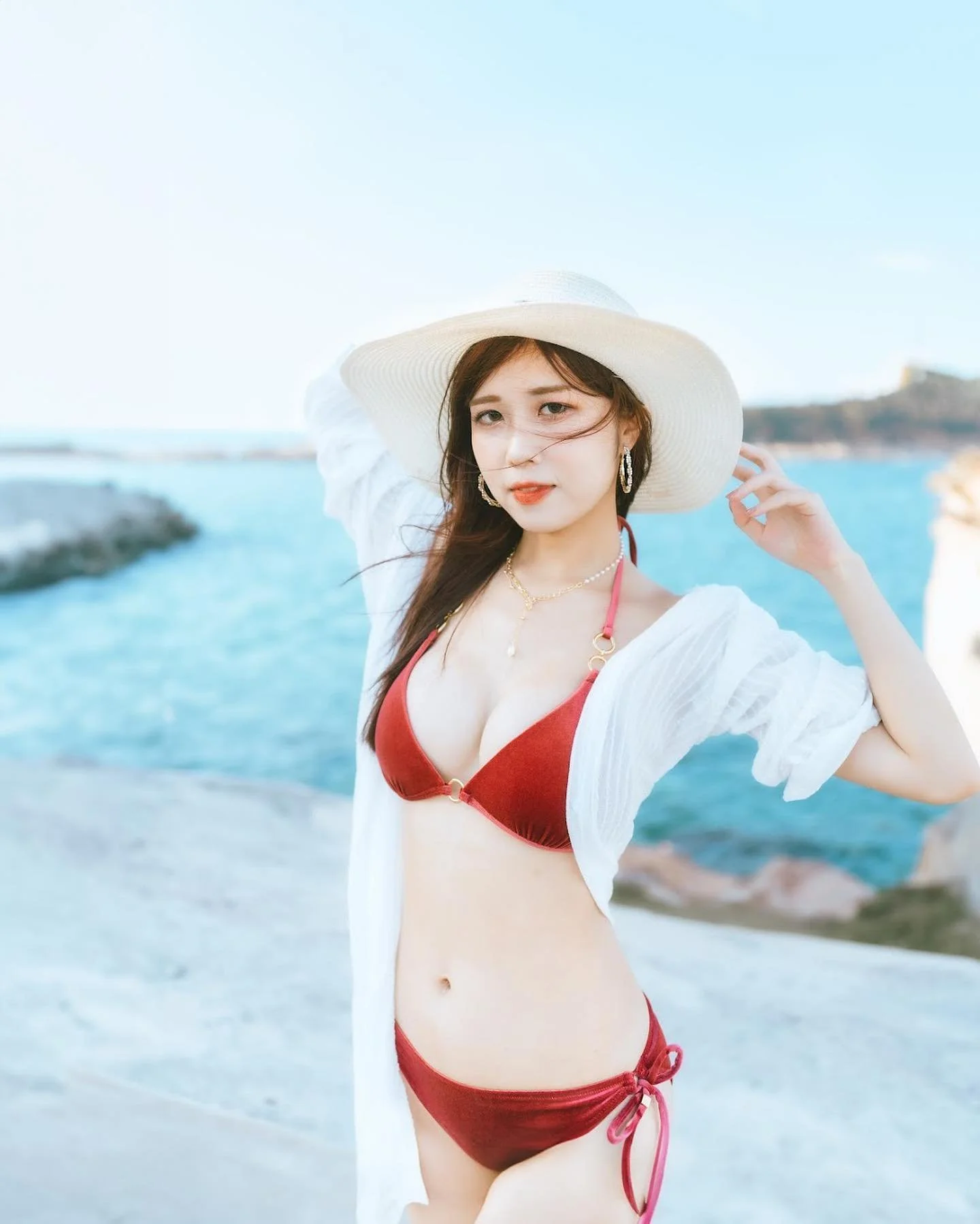 一名女性穿著紅色比基尼和白色外套，站在海邊並微笑，背後是碧藍的海水和晴朗的天空。