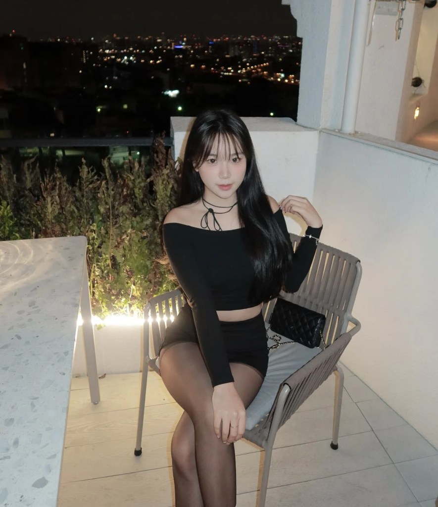 一位女性坐在椅子上，穿著黑色上衣和短裙，背景是夜晚的城市燈光。