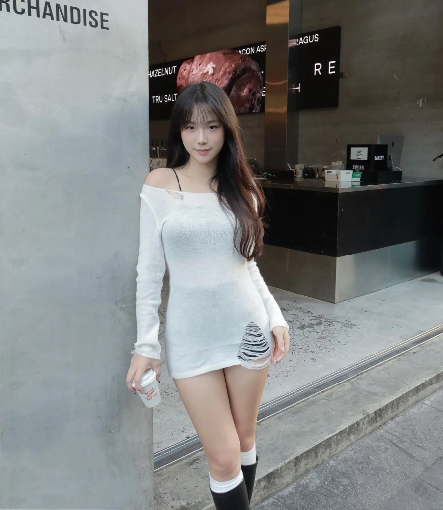 一位女性穿著白色毛衣裙，搭配黑色過膝襪，微笑站在商店前，手裡拿著飲料。