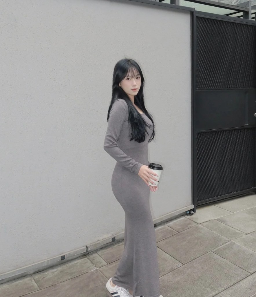 一位女性穿著灰色長袖連衣裙，手持咖啡杯，站在室外背景前