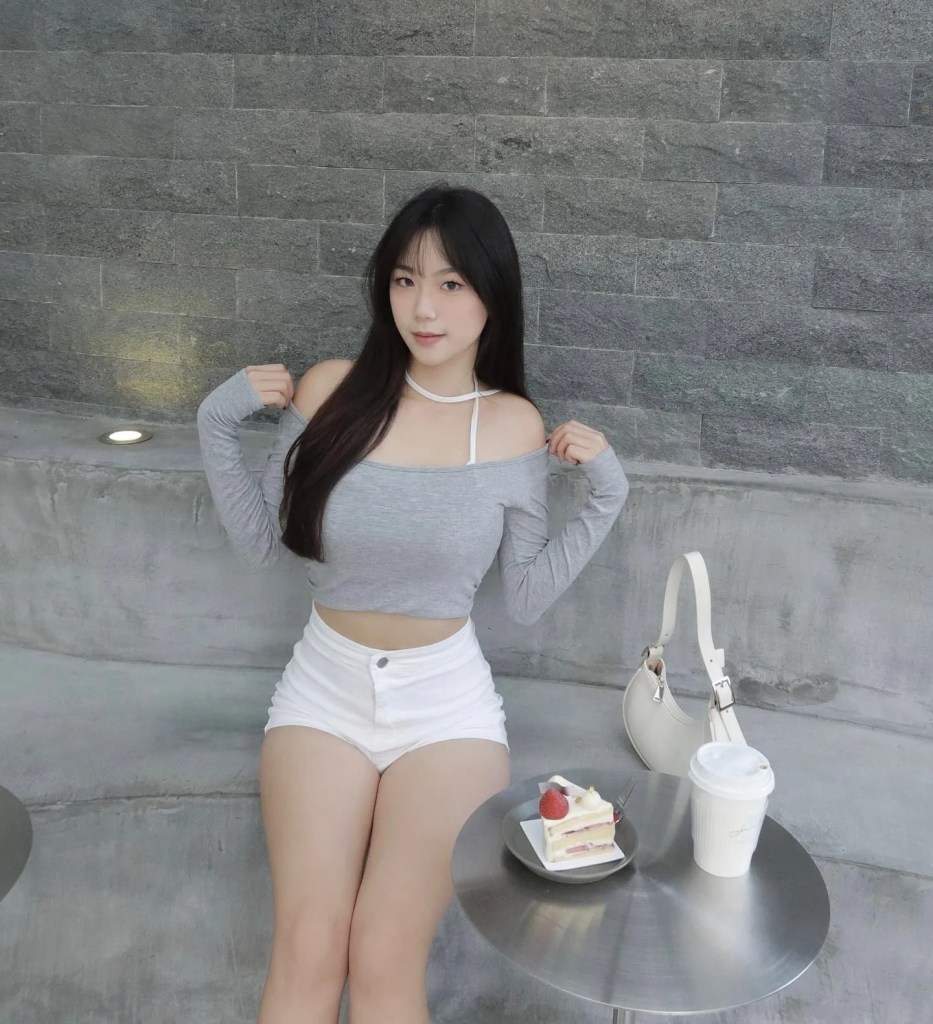一位穿著灰色露肩上衣和白色短褲的年輕女性坐在咖啡桌旁，旁邊有蛋糕和咖啡，背景是灰色石牆。