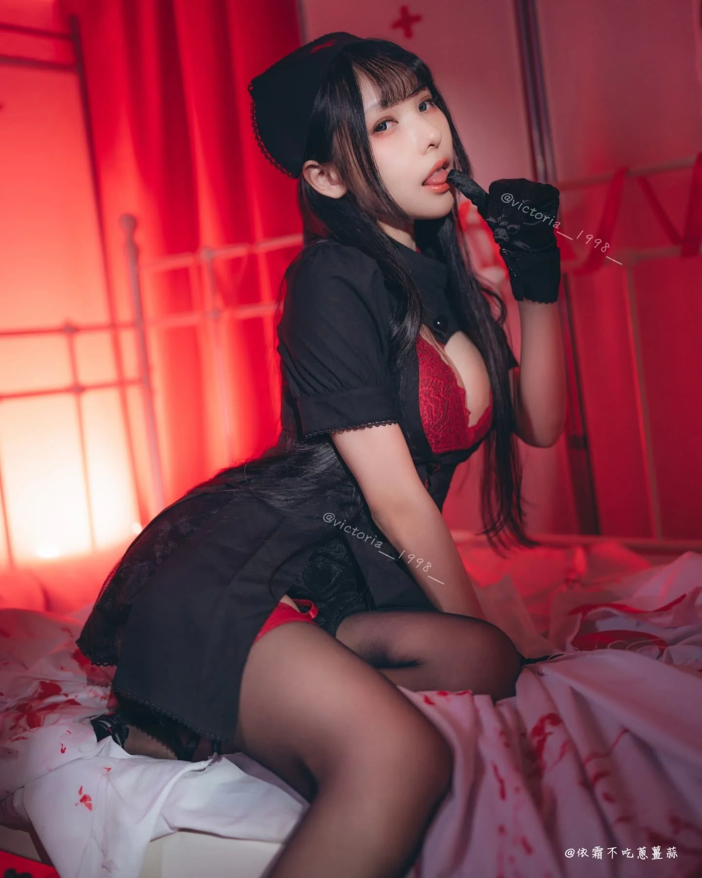 一位穿著黑色護理服的女性坐在紅色燈光下的床上，表情性感地舔著手指，背景帶有血紅色的裝飾，營造出神秘氛圍。