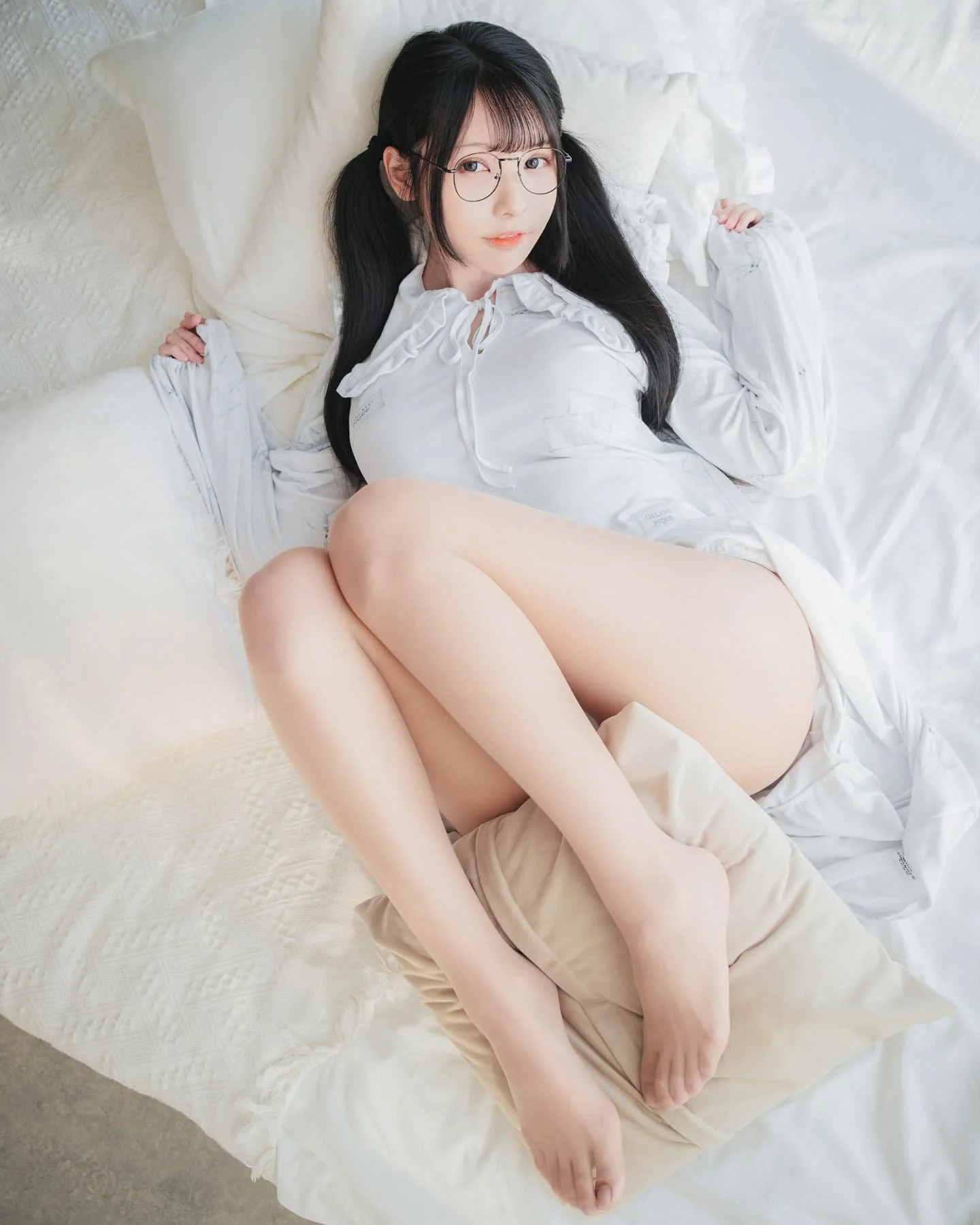 一位女孩穿著白色上衣，坐在床上，雙手撐著身體，腿交疊著，周圍有柔軟的枕頭和床單。