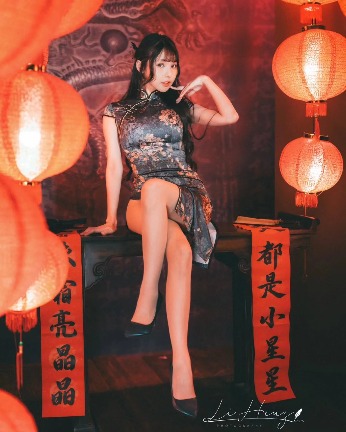 一位穿著旗袍的女性坐在桌子上，手指著臉，背景有紅色燈籠和裝飾，展現出節日慶祝的氛圍。