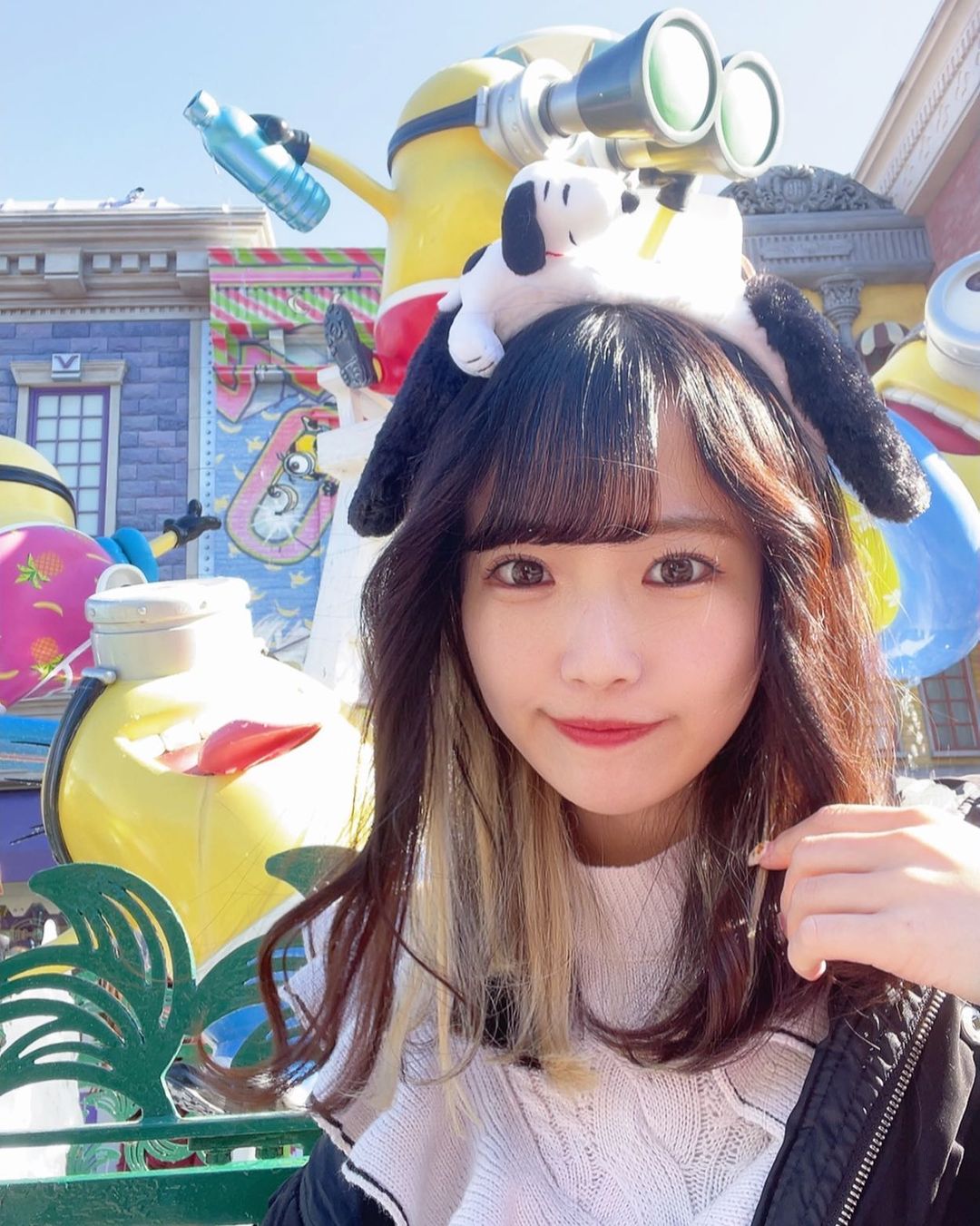 一名女性在主題公園前拍照，頭上戴著一隻小狗造型的帽子，背景有色彩繽紛的裝飾和玩具。