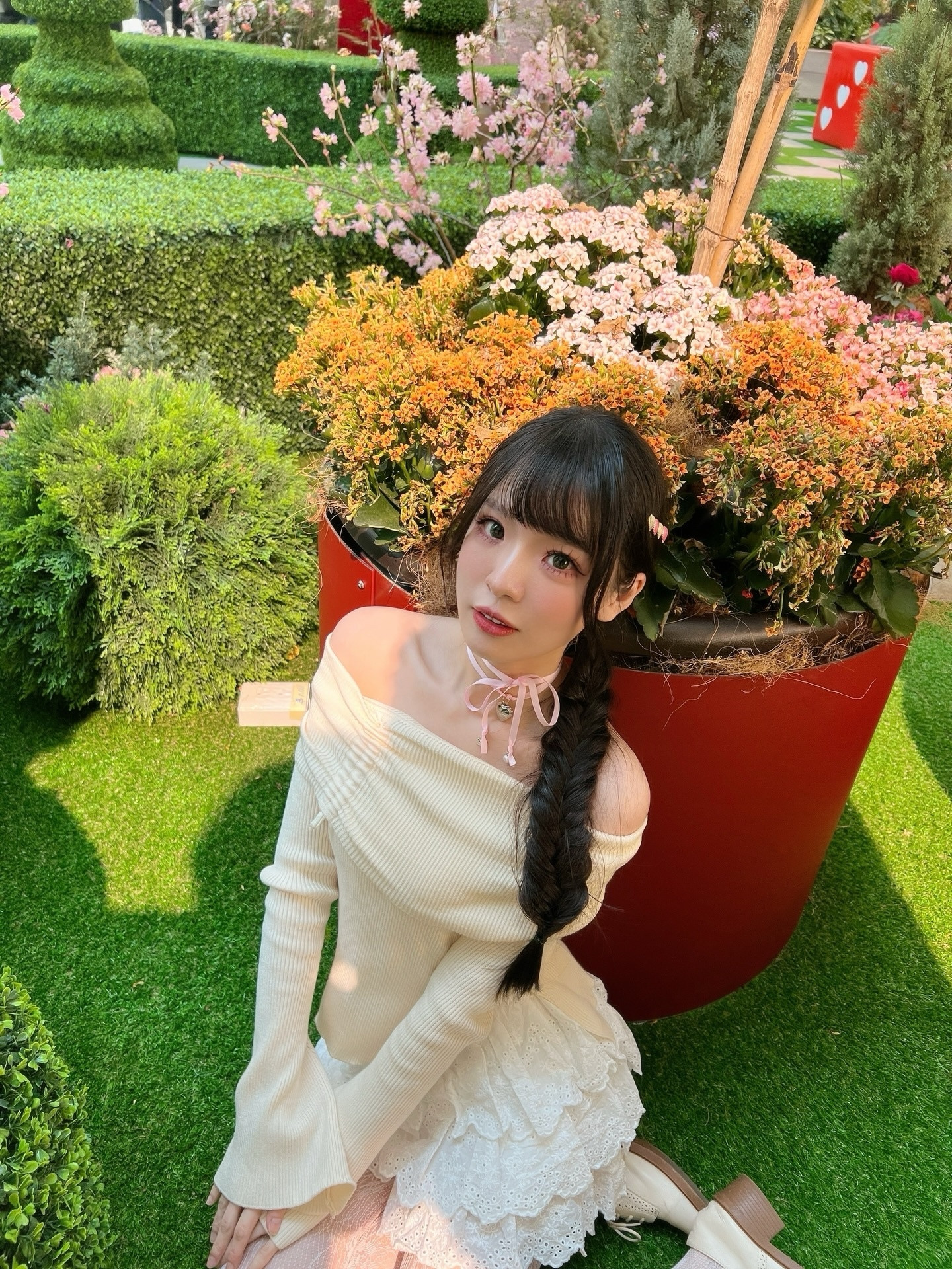 一位穿著淺色毛衣和白色裙子的年輕女性，坐在多彩花卉和綠色植物前，背景中有藝術造型的植物和紅色花盆。