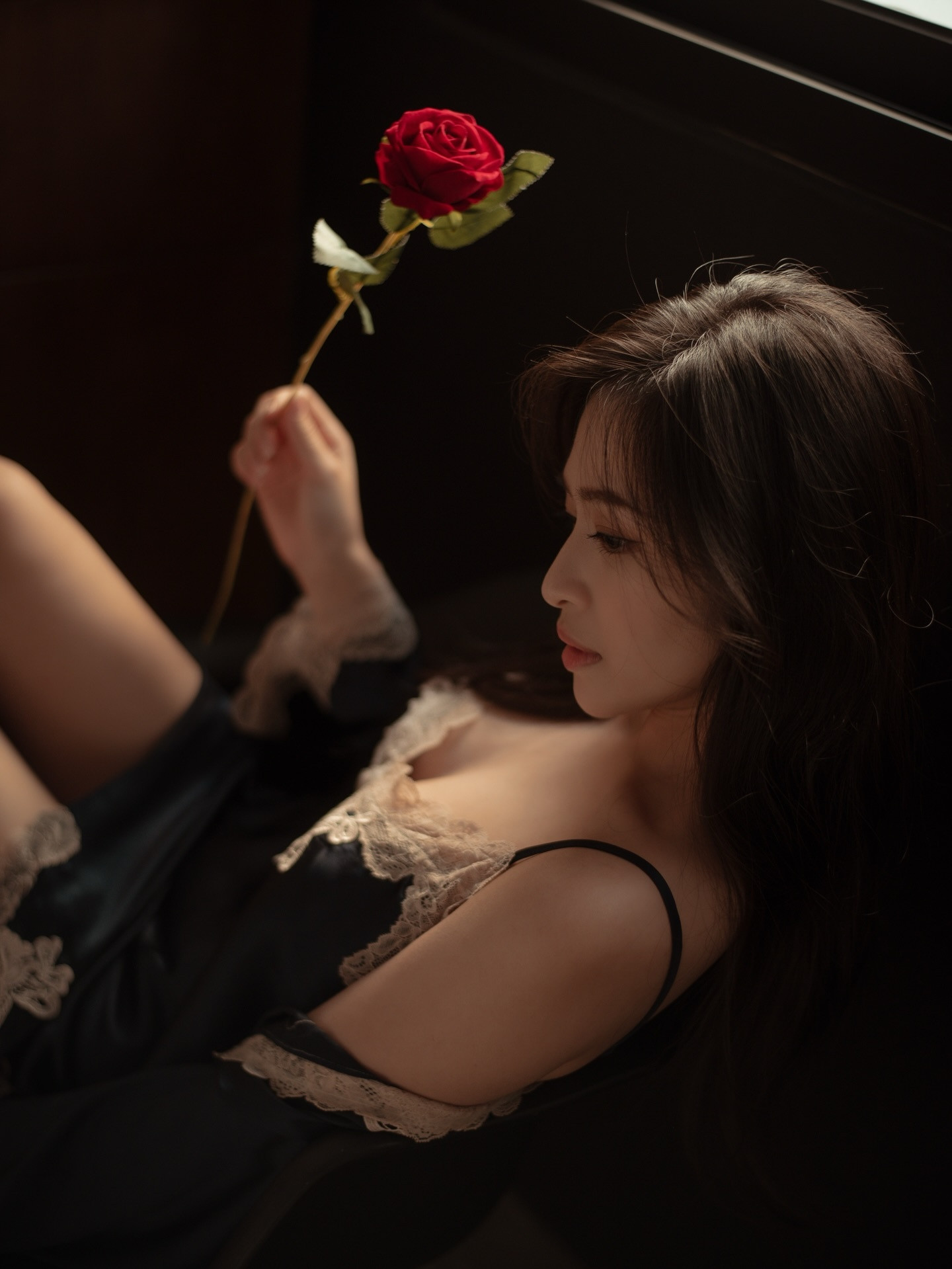 一位女子手持紅玫瑰，身穿淺色蕾絲邊的深色睡衣，坐在窗邊，臉部側影展現出優雅的姿態。