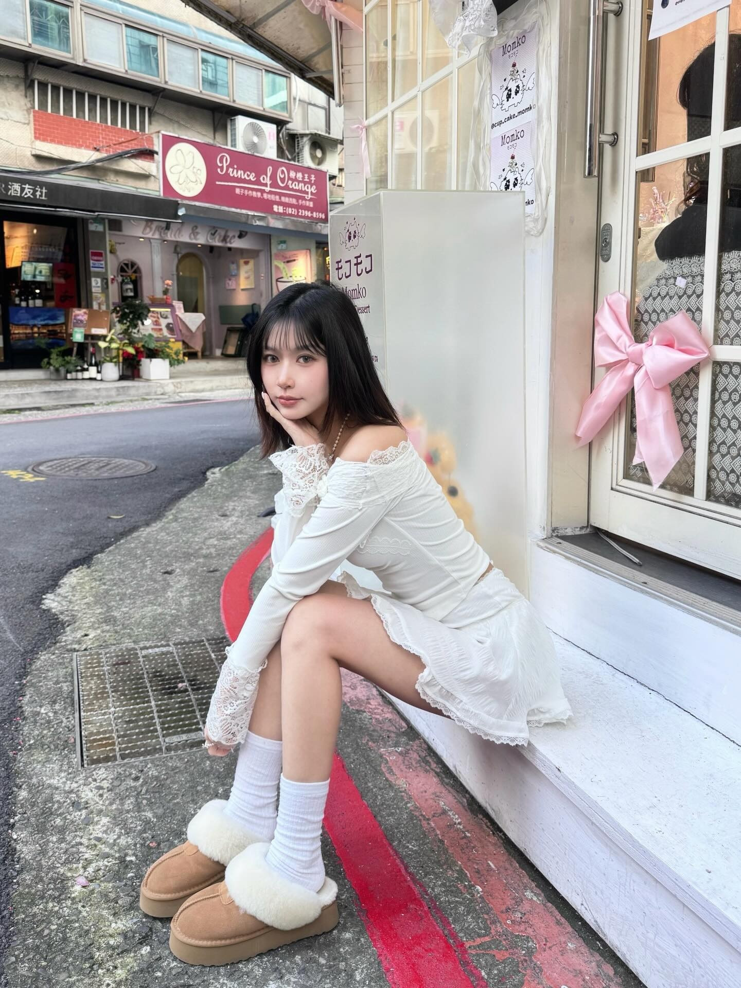 一名女子穿著白色上衣和裙子，坐在街道旁的白色牆邊，腳穿毛絨拖鞋。背景是一家商店，周圍環境充滿城市街道氣息。