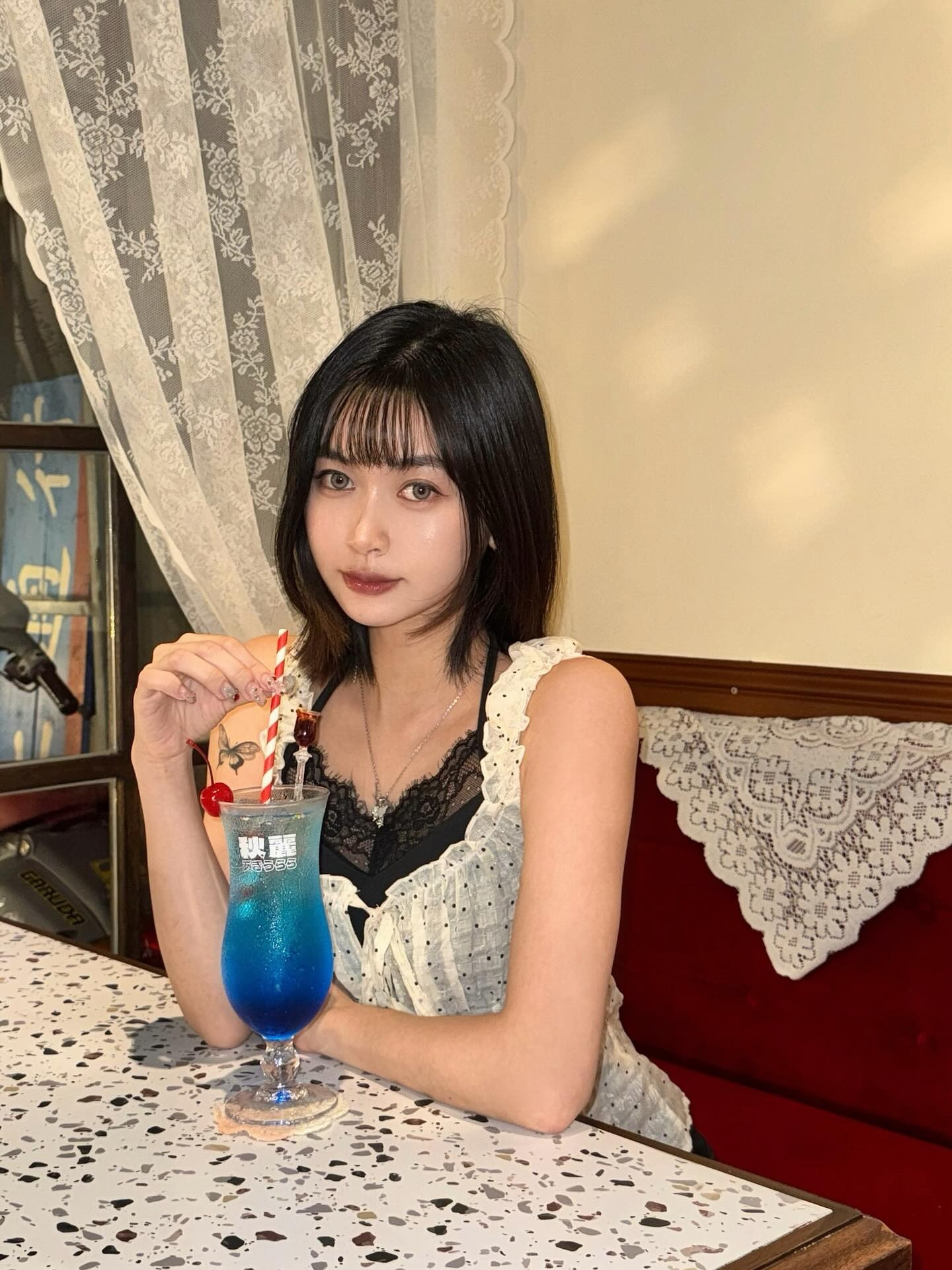 一位年輕女性坐在桌子旁，手中拿著一杯藍色的飲品，上面裝飾著櫻桃和吸管，背景有蕾絲窗簾和紅色沙發。