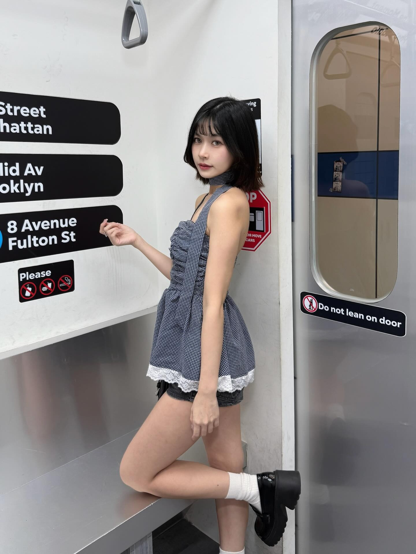 一位年輕女性在地鐵車廂內，穿著格子上衣和短褲，靠在金屬長椅上，背景有地鐵路線指示牌。