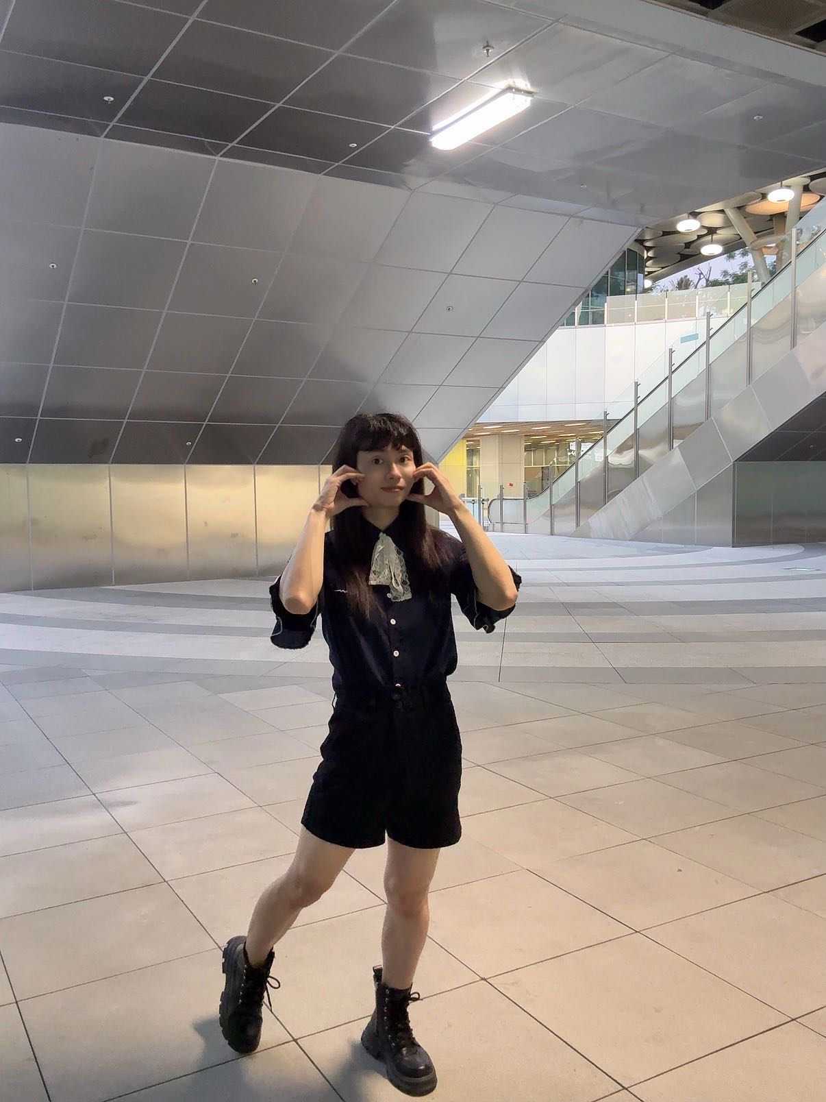 一位女孩在現代建築內微笑並比心，穿著黑色襯衫和短褲，搭配黑色靴子。