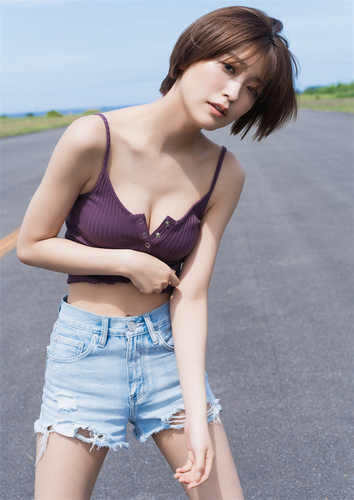 一名女性穿著紫色無袖上衣和淺藍色破洞短褲，站在公路上，背景是晴朗的天空和綠色的景色。