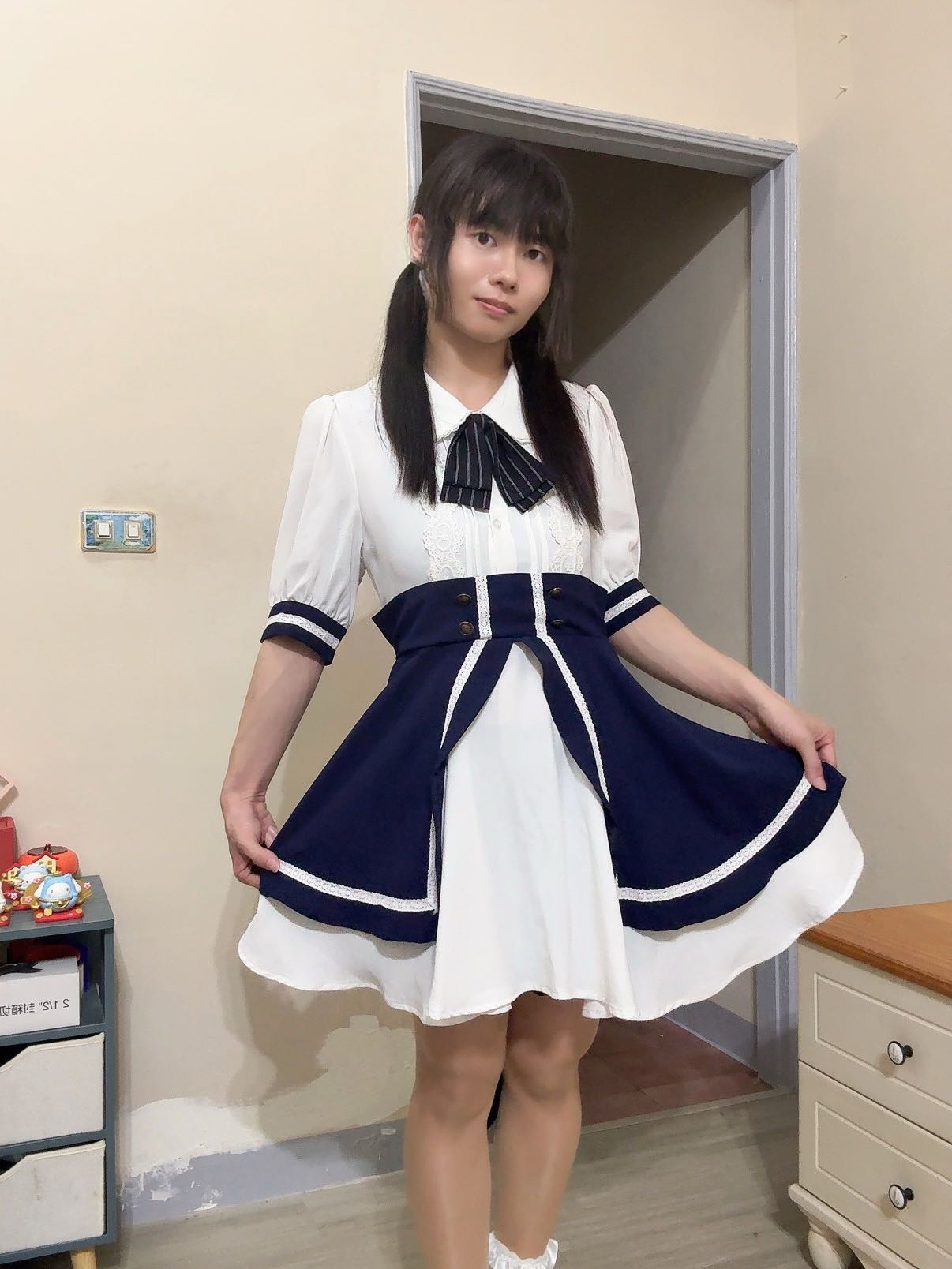 一位穿著白色和深藍色連衣裙的女性，站在房間內微笑，裙子展開，搭配領帶，背景有家具和牆壁。