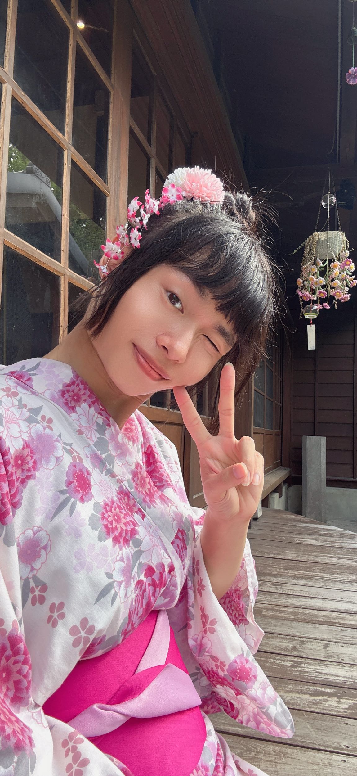一位穿著粉紅花紋和服的女性，坐在木質平台上，微笑並做出勝利手勢，背景有窗戶和吊飾。