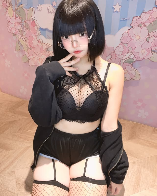 一位穿著黑色蕾絲內衣和短褲的女性，坐在背景為粉色花卉圖案的地方，手指輕觸下巴，配戴眼鏡，表情甜美。