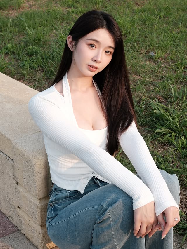 一名年輕女性坐在草地邊緣，穿著白色長袖上衣和藍色牛仔褲，面帶微笑，目光柔和。