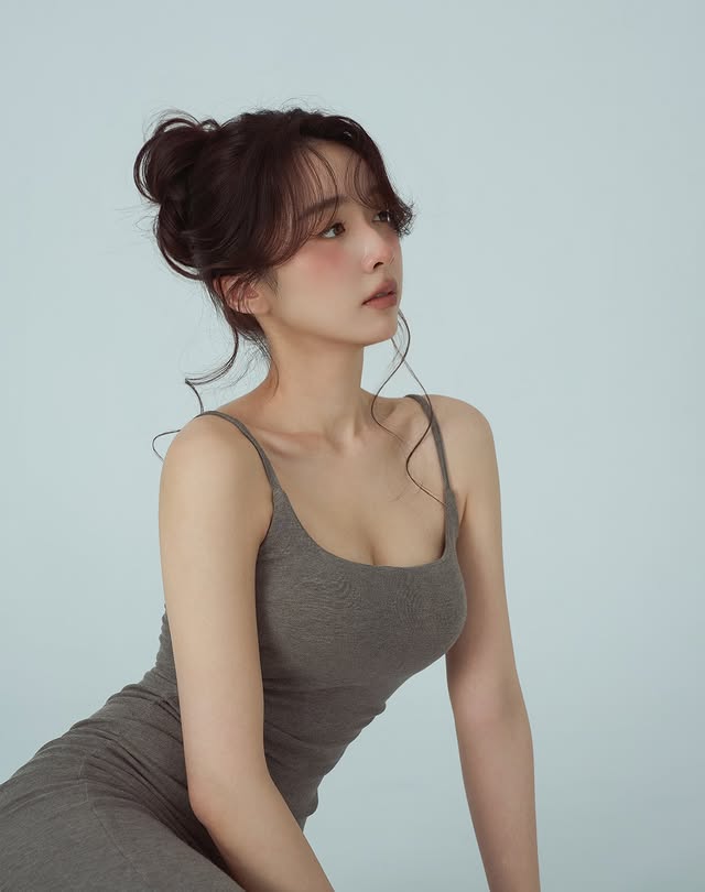 一位年輕女性側身坐著，穿著灰色的無袖連衣裙，頭髮蓬鬆卷曲，背景為柔和的淺藍色。