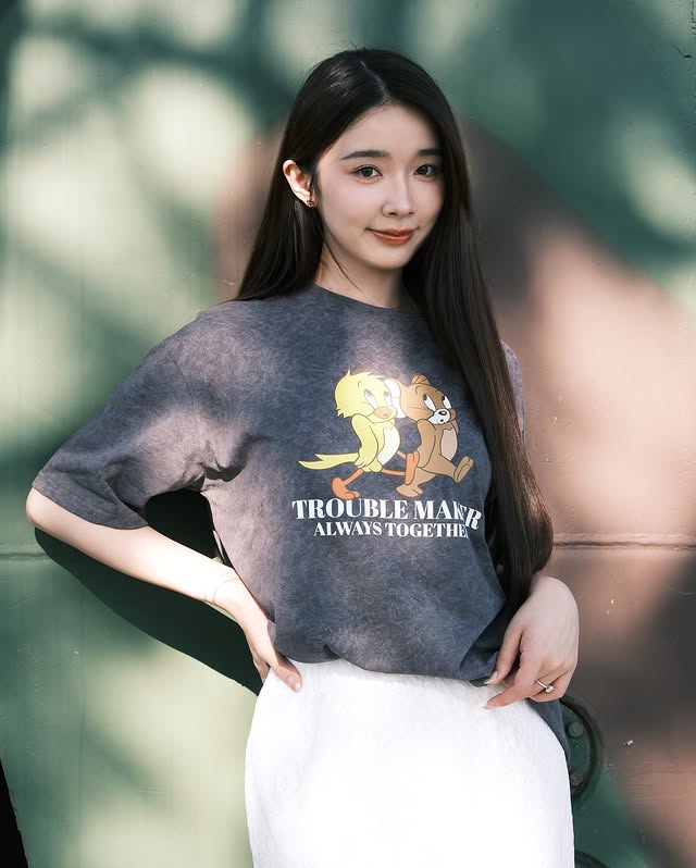 一位女性站在綠色牆前，穿著淺色T恤，印有卡通角色和字樣 'TROUBLE MAKER ALWAYS TOGETHER'，髮長飄逸，表情親切。