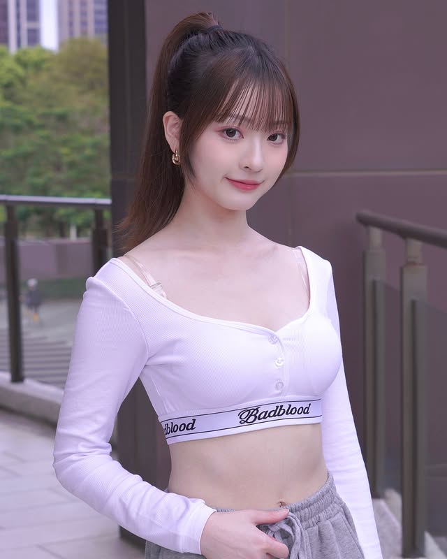 一位年輕女性穿著白色長袖上衣和灰色運動褲，微笑著站在戶外，背景有綠樹和現代建築。