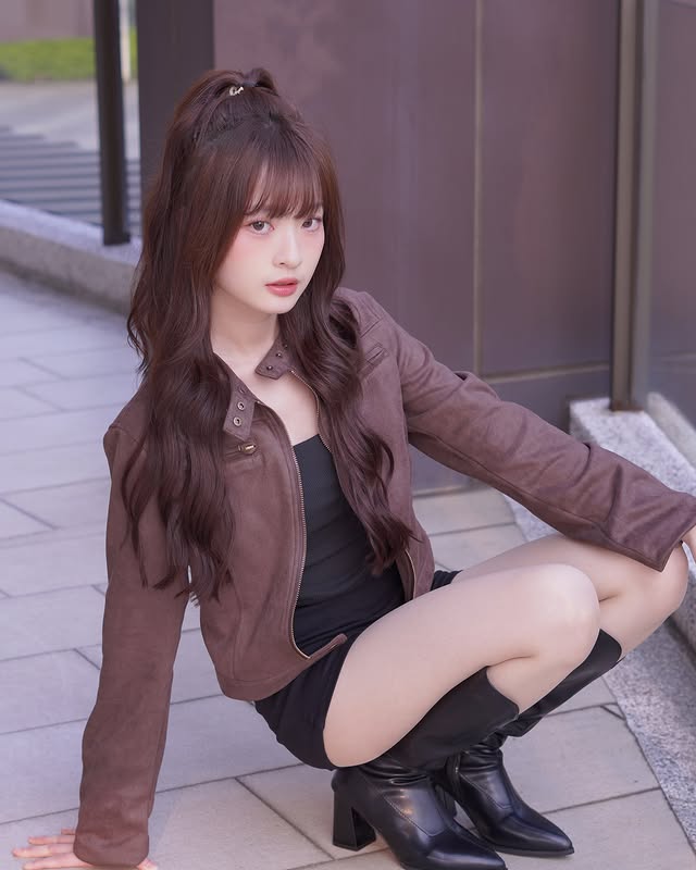 一位女性坐在街道上，穿著棕色外套和黑色靴子，長髮披肩，面對鏡頭展現自信的姿態。