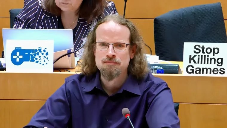一名男子坐在會議桌前,表情嚴肅,穿著深色襯衫,桌上有帶有遊戲符號的標誌和寫著"Stop Killing Games"的牌子,背景中有另一名女性參與討論。