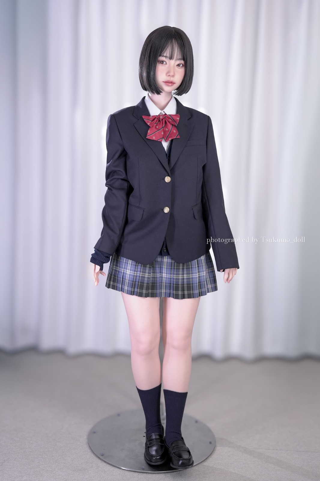 一位穿著校服的女孩,身穿深色外套和格子裙,搭配紅色領結,展示在白色背景前。