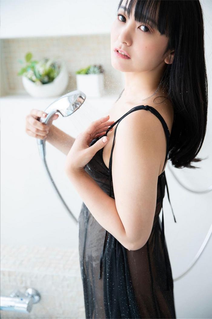 一名女性站在淋浴間，穿著透明的黑色衣物，手持淋浴槍，面向鏡頭，表情柔和，背景有植物裝飾。