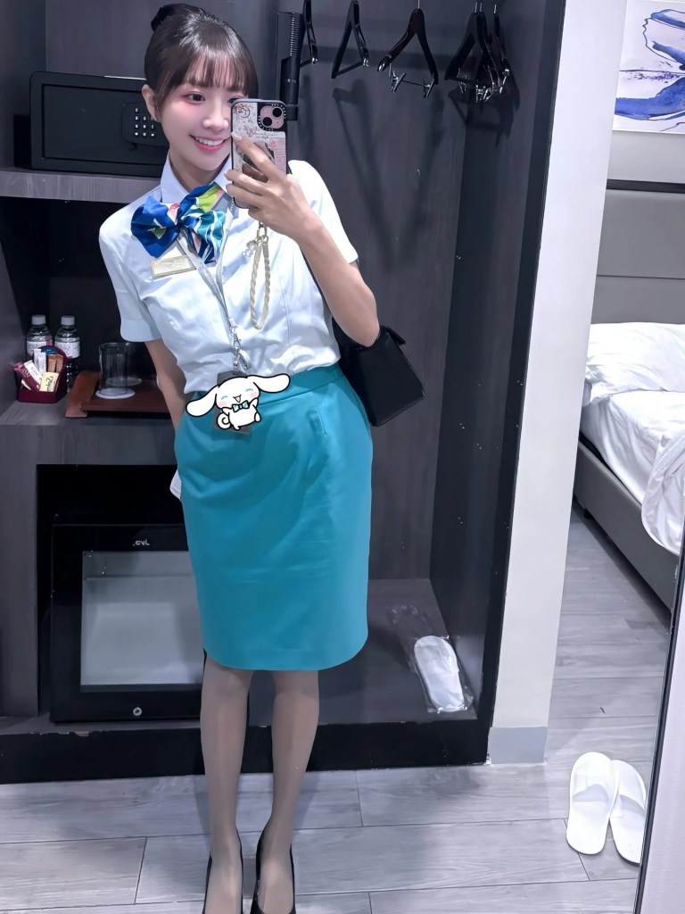 一位女性穿著制服，在鏡子前自拍，搭配彩色圍巾和藍色裙子，背著黑色手提包，房間內有衣架和床。