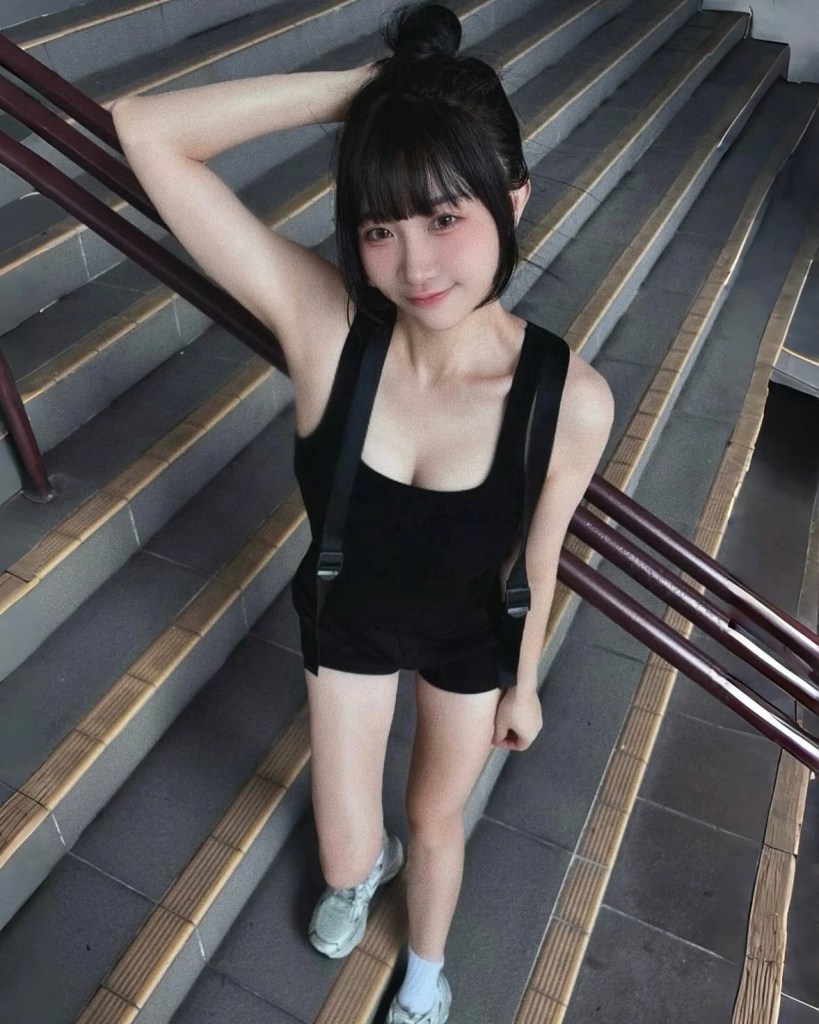 一位穿著黑色背心和短褲的年輕女性，站在樓梯上，微笑並把手撐在頭上。