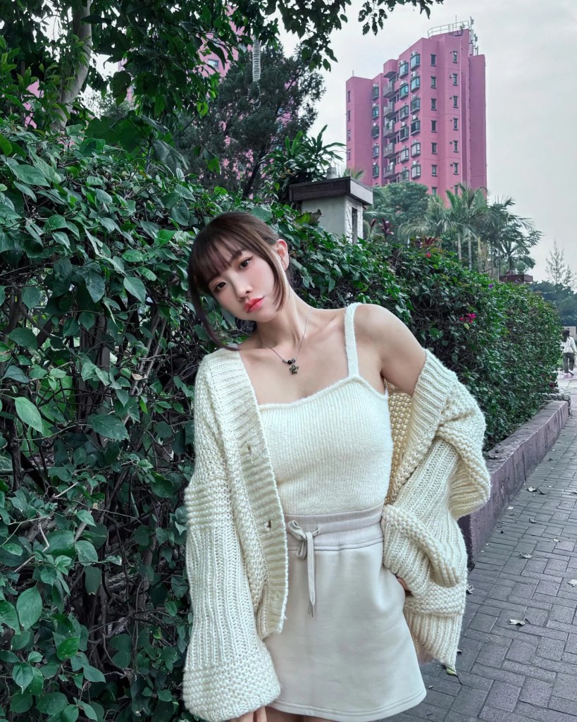 一名女性穿著白色毛衣和裙子，站在綠色植物旁邊，背景有粉紅色的大樓。