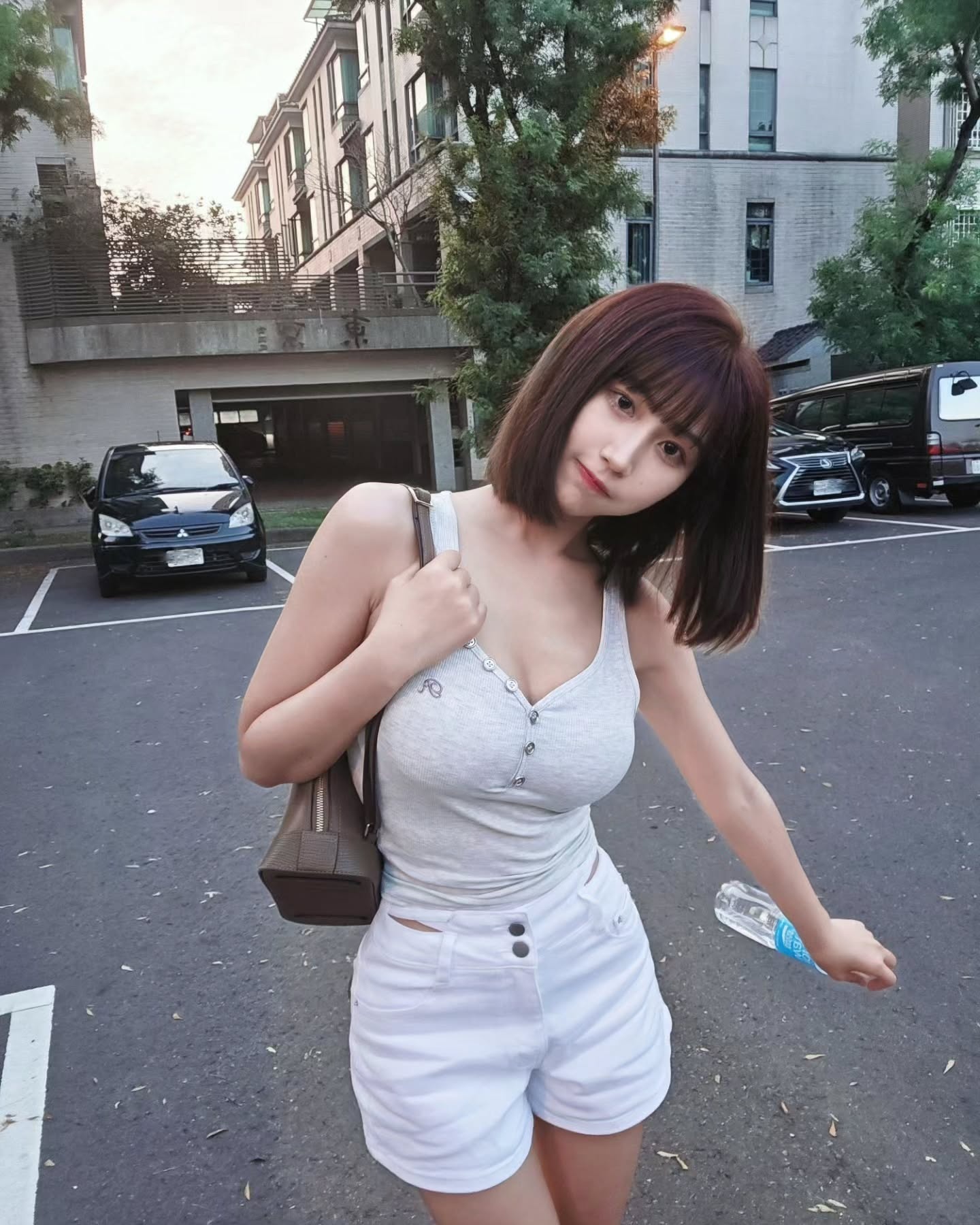 一名女性在停車場中站立，穿著灰色背心和白色短褲，手拿水瓶，背景為城市建築和樹木。