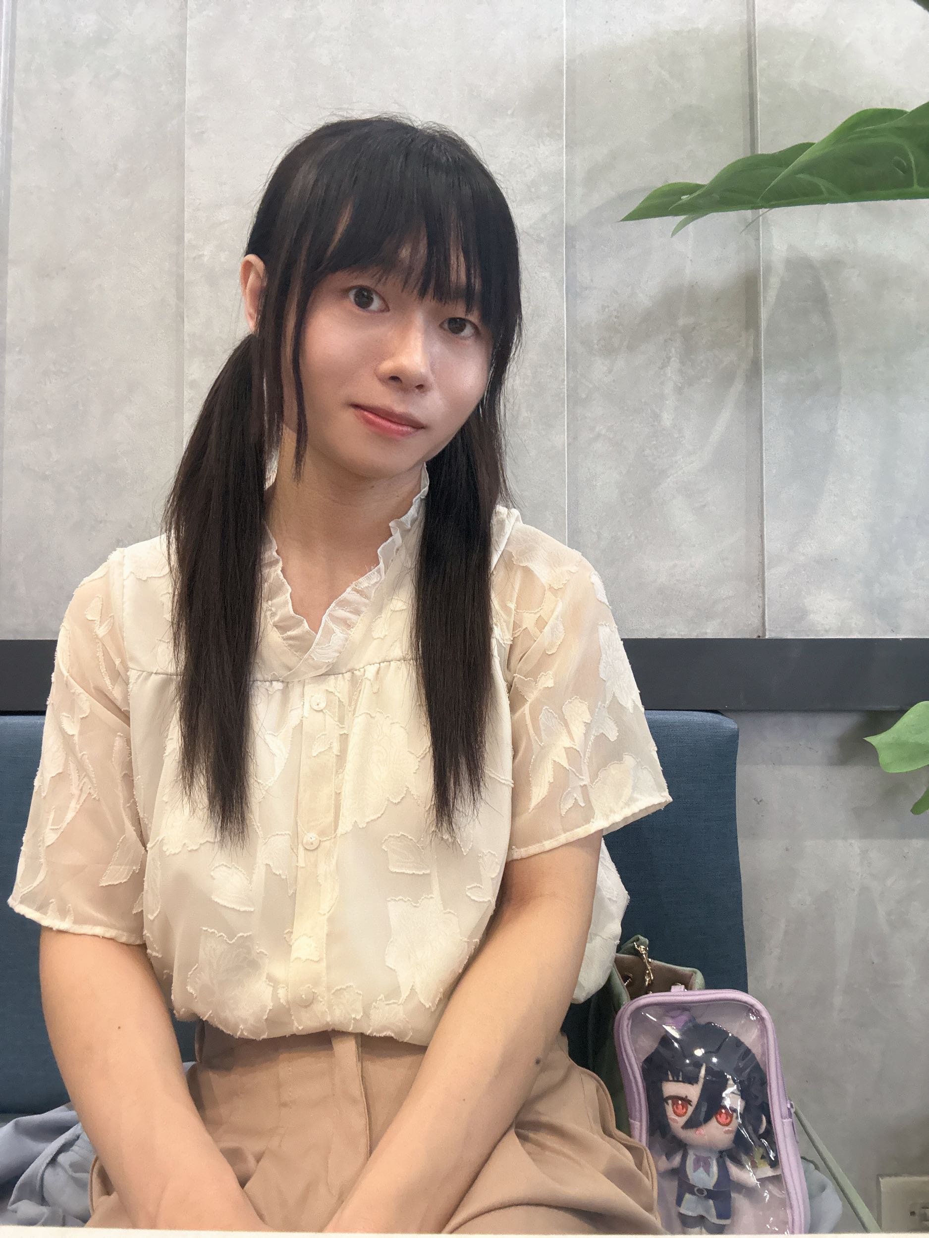 一名年輕女性坐在沙發上，穿著淺色襯衫和淺色褲子，身旁放著一個裝有人物造型的透明包包，背景有綠色植物和淺色牆面。