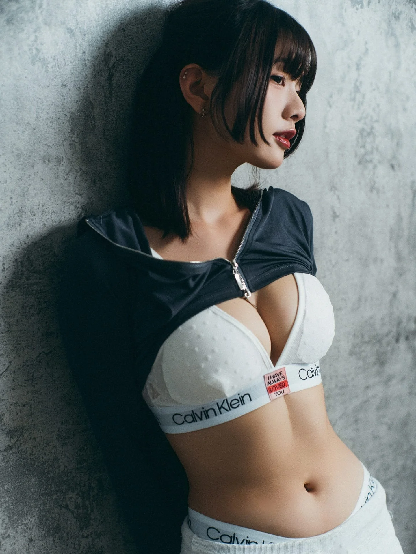 一位女性側身靠在混凝土牆上，穿著黑色夾克和白色Calvin Klein內衣，表情柔和。