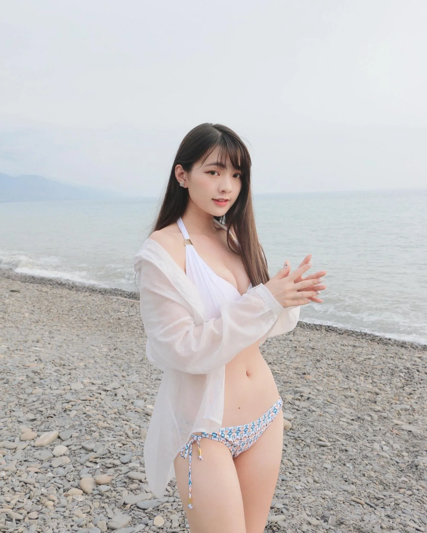 一位年輕女性穿著比基尼和輕薄外套，站在海邊的卵石上，手指在一起，背景是平靜的水面和灰色的天空。