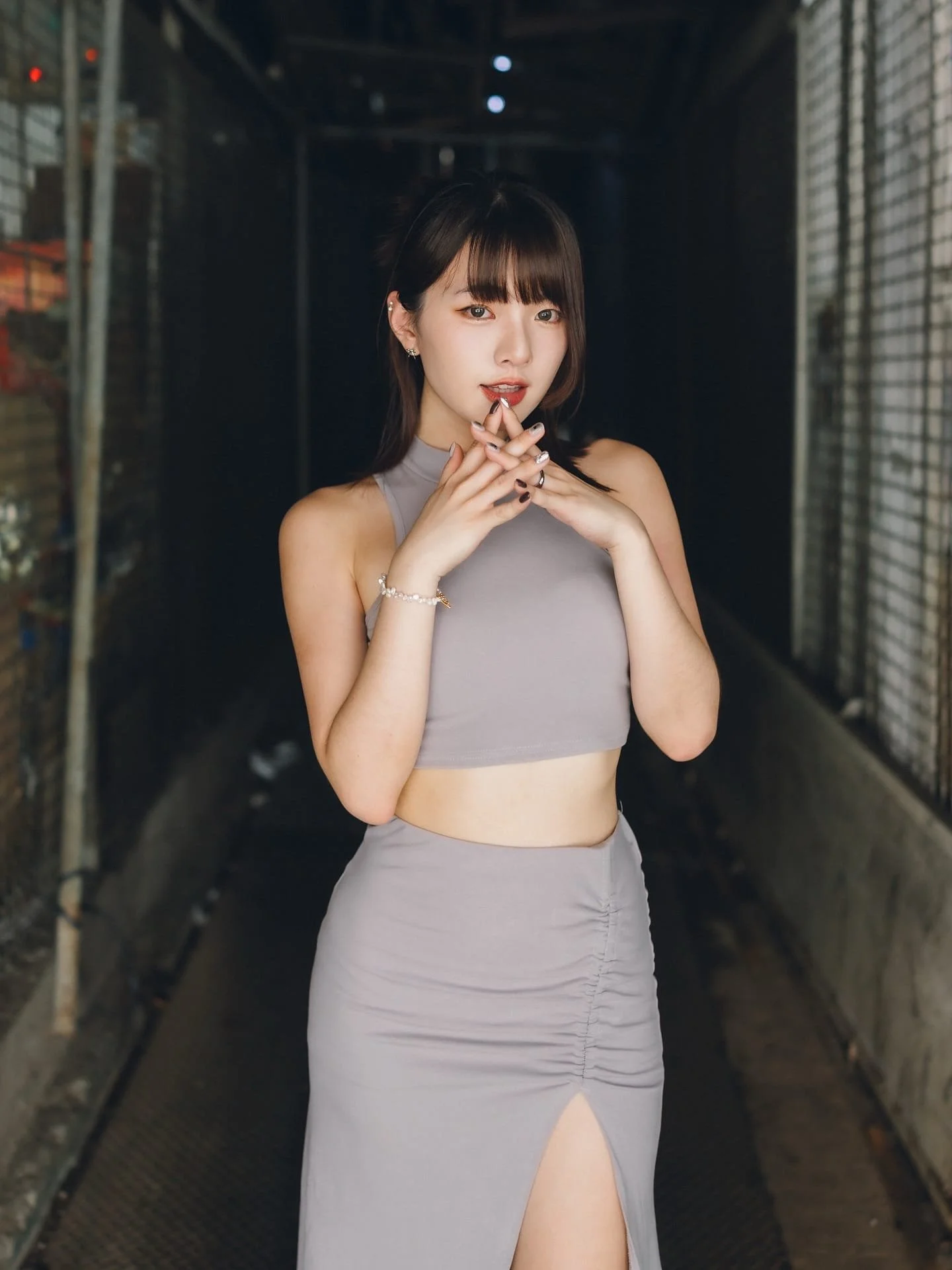 一位女性穿著灰色緊身上衣和裙子，站在狹窄的走廊中，手指交叉在唇邊，目光凝視鏡頭。