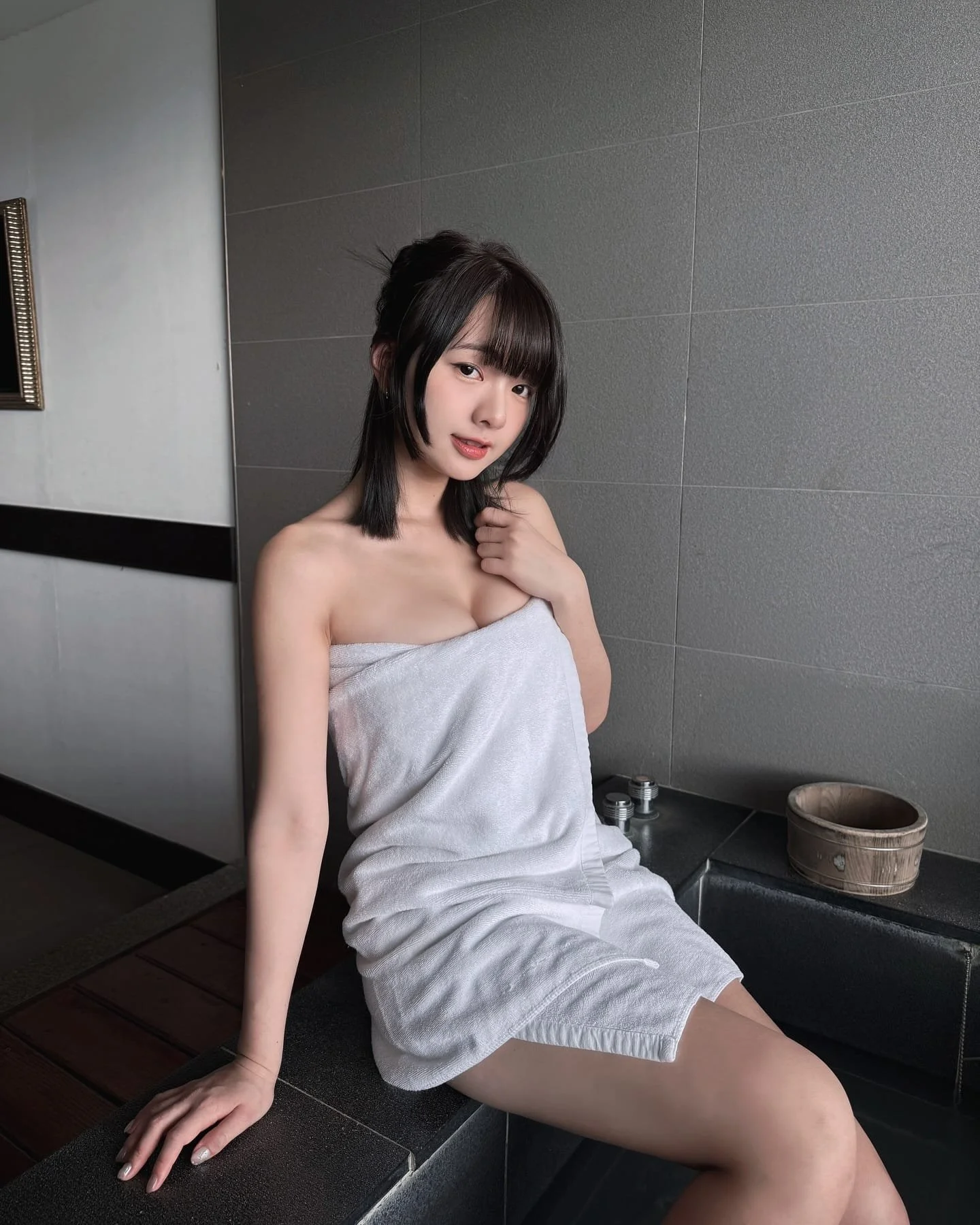 一位年輕女性坐在浴缸邊，身穿白色毛巾，微笑著看向鏡頭，背景為簡約的室內浴室設計。