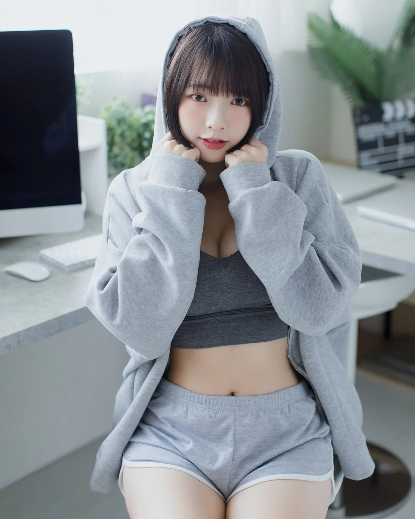 一位穿著灰色帽衫和短褲的女性坐在桌前，面帶微笑，雙手托住帽衫的前襟。背景有電腦和植物，環境明亮舒適。