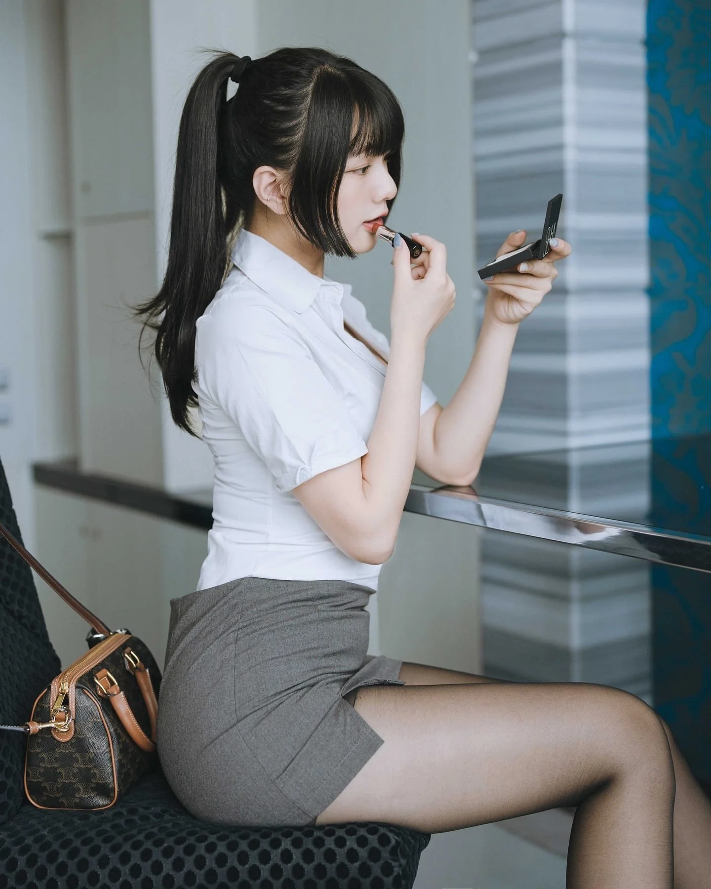 一位女士坐在椅子上，穿著白色襯衫和灰色裙子，正在用小鏡子塗口紅。