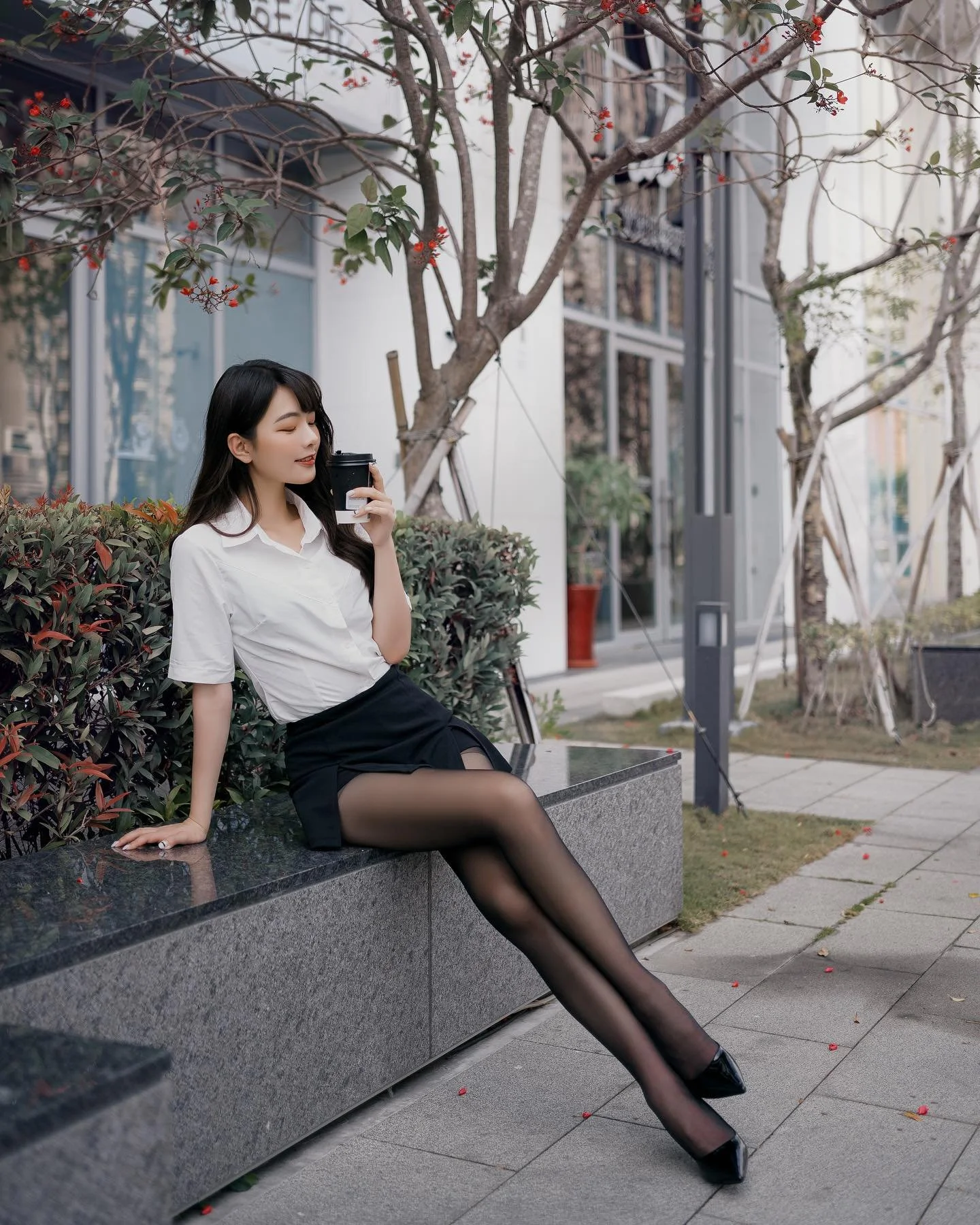 一位穿著白色襯衫和黑色短裙的女性坐在石凳上，手中握著咖啡，背景有樹木和商店。
