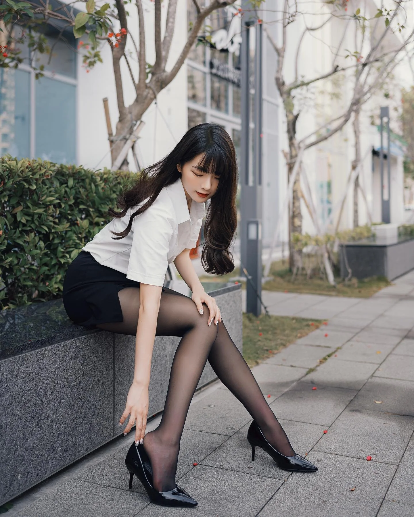 一位女性穿著白襯衫和黑色短裙，坐在石墩上，調整著她的高跟鞋，周圍有綠色植物和城市背景。